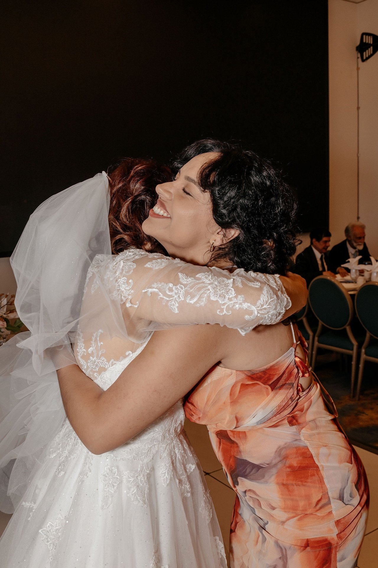 The bride, in a lace gown and long veil, hugs a smiling guest in a peach floral dress at the reception