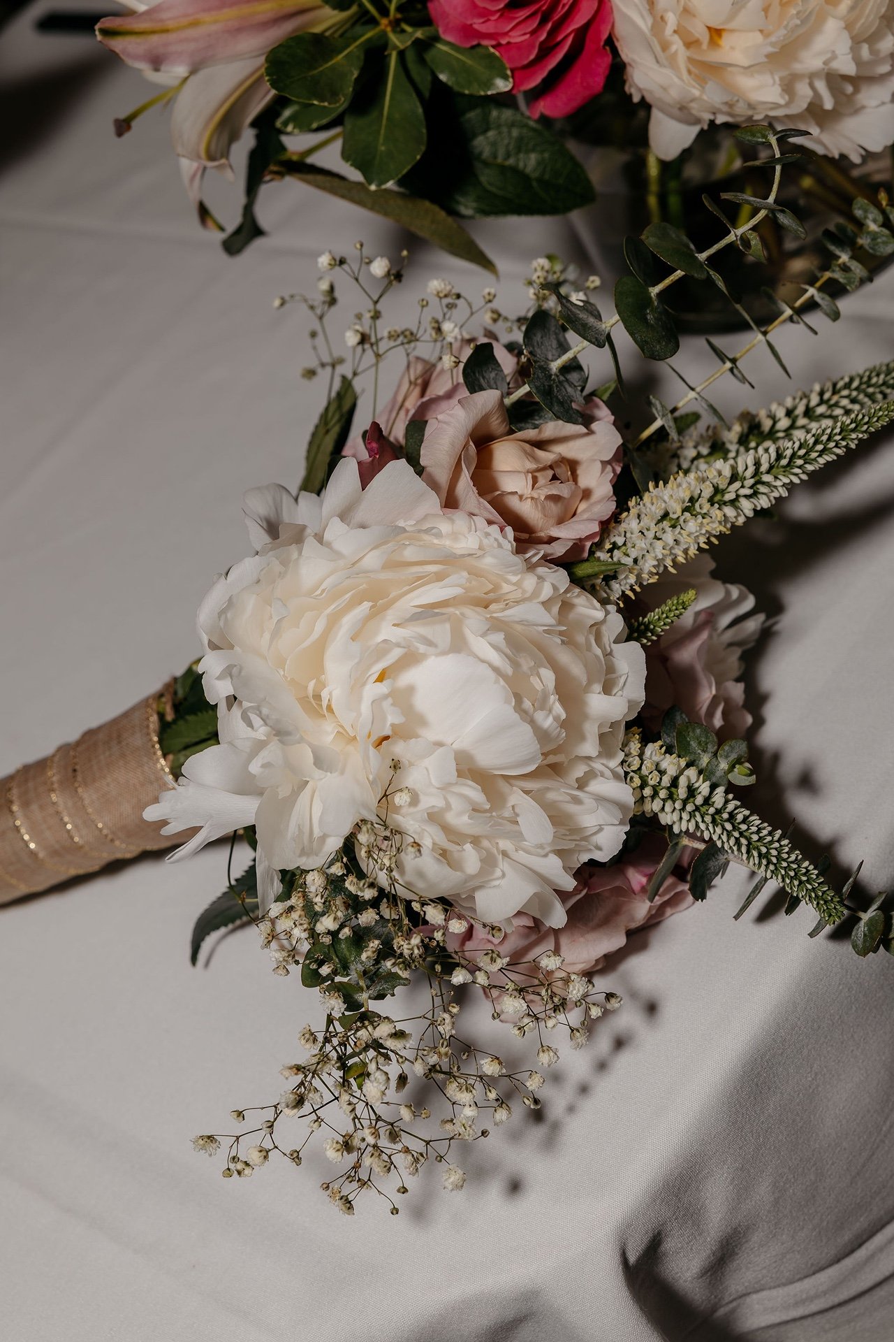 A bridal bouquet of a white peony, pale pink roses and baby's breath, its stems wrapped in gold-striped ribbon, resting on white table linen