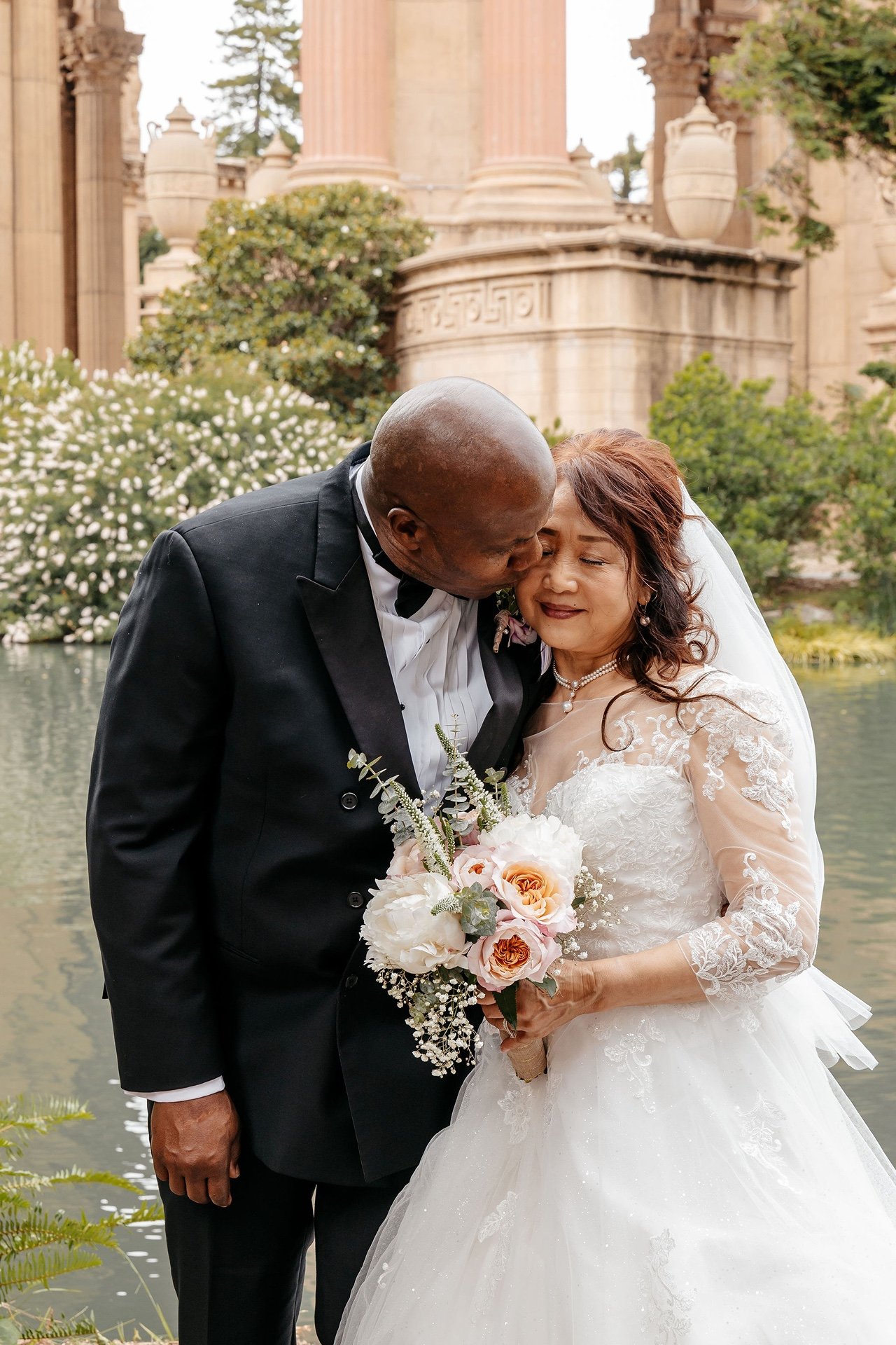 The groom kisses the bride on the cheek beside the lagoon at the Palace of Fine Arts, her bouquet of peonies and roses held between them