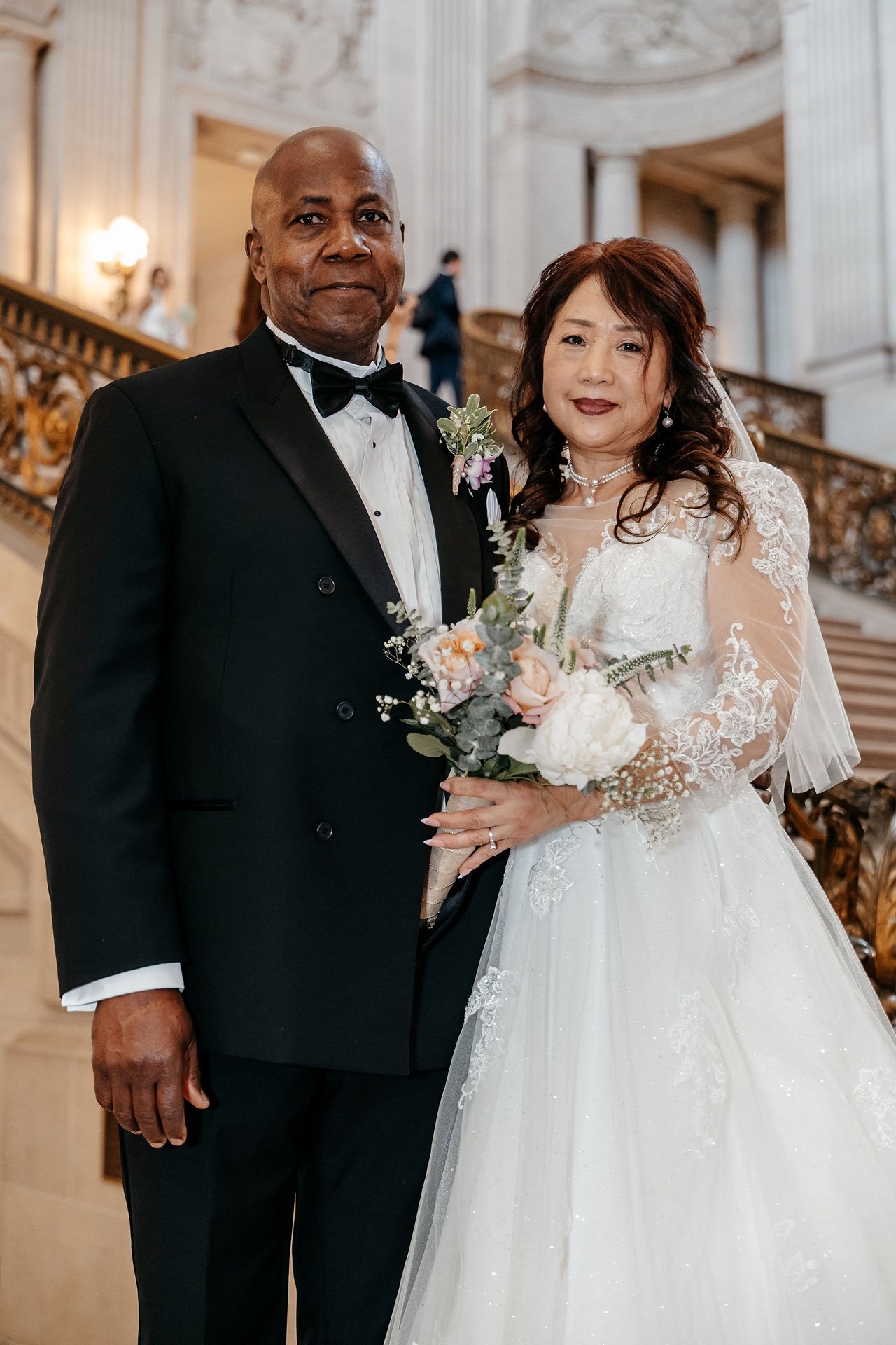 A groom in a black double-breasted tuxedo and a bride in a lace gown holding peonies and roses, posed on the grand staircase at San Francisco City Hall