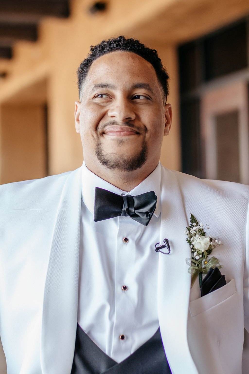 A groom wearing a white tuxedo and black bow tie smiles and thinks about questions to ask your wedding photographer