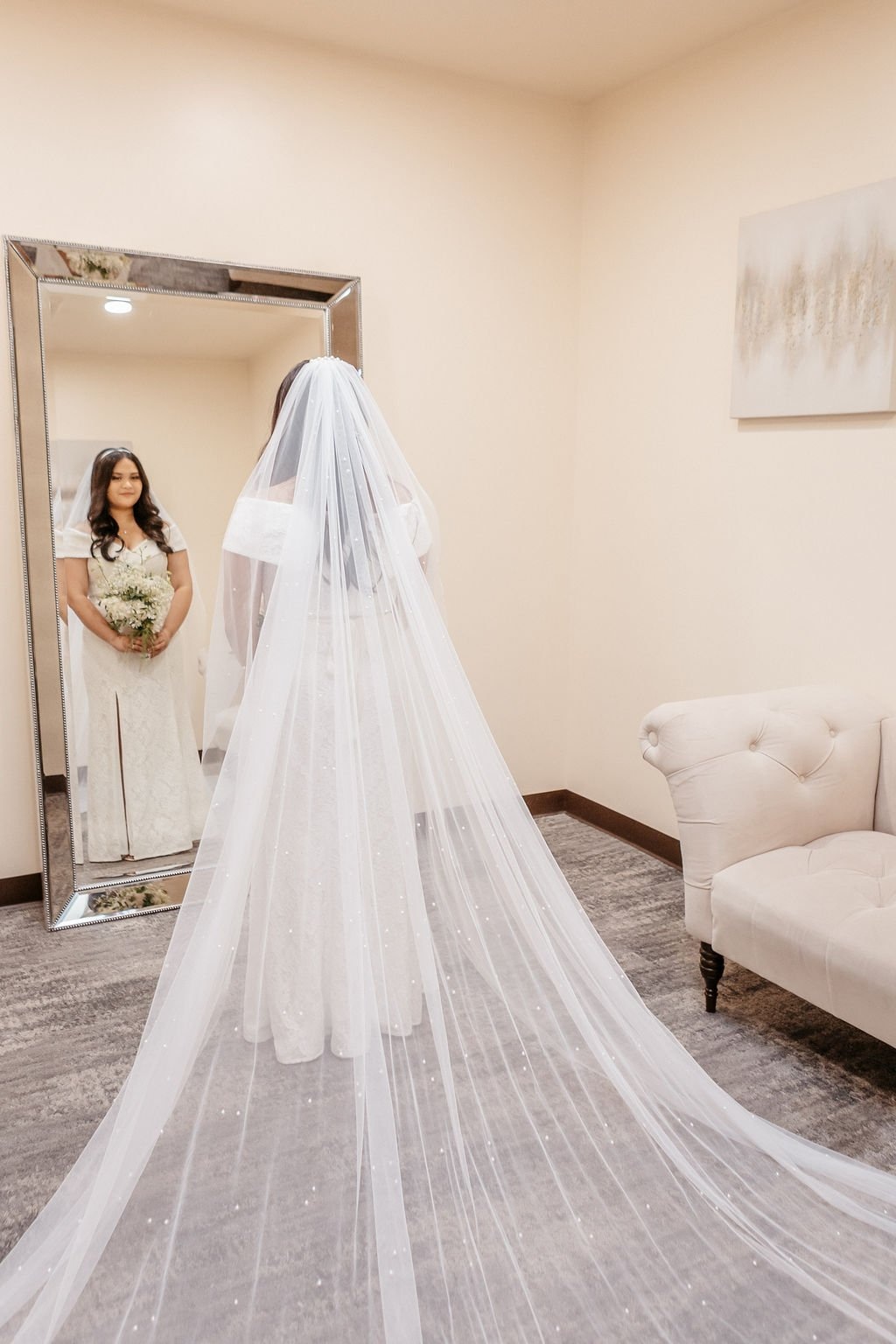 A bride with a long white veil looks into the mirror while holding flowers knowing what questions to ask your wedding photographer