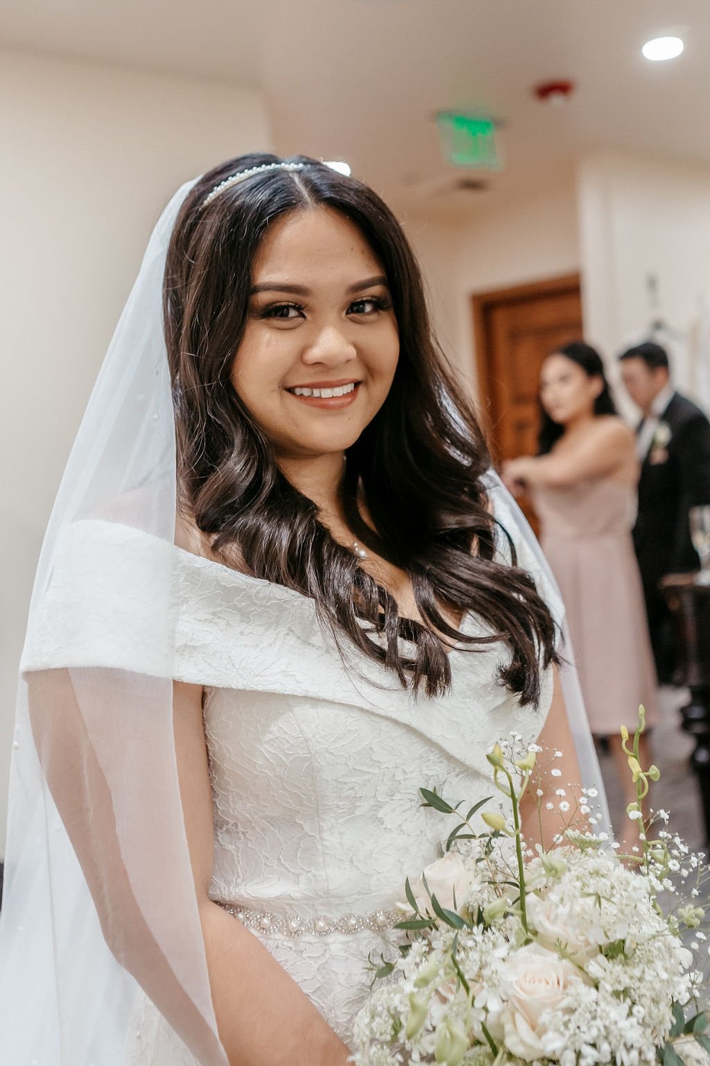 A bride with dark brown hair smiles while wearing her wedding dress and veil holding white flowers knows what questions to ask your wedding photographer before booking