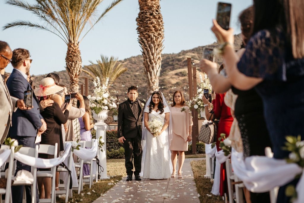 A bride and her mother and father walk down the aisle towards her groom while a photographer takes sf wedding photos